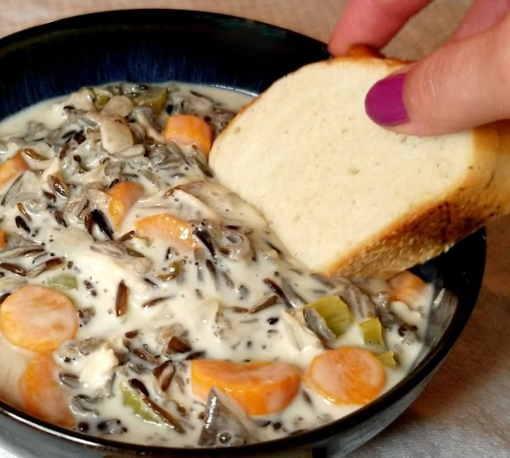 a person is dipping some bread into a bowl of chicken and wild rice soup with carrots