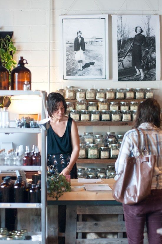 two women standing in front of a counter with jars on the wall and pictures above it