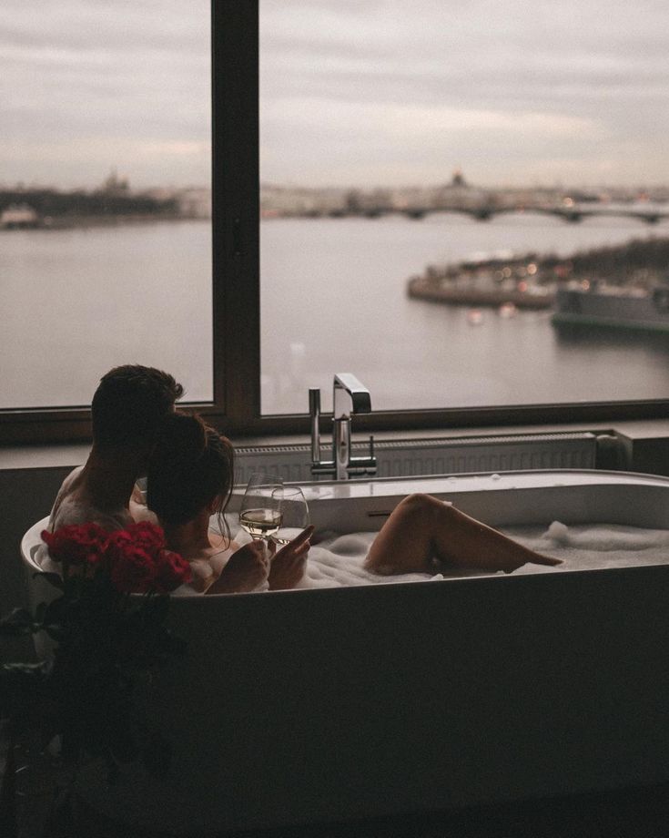 two people sitting in a bathtub with a view of the water and boats outside