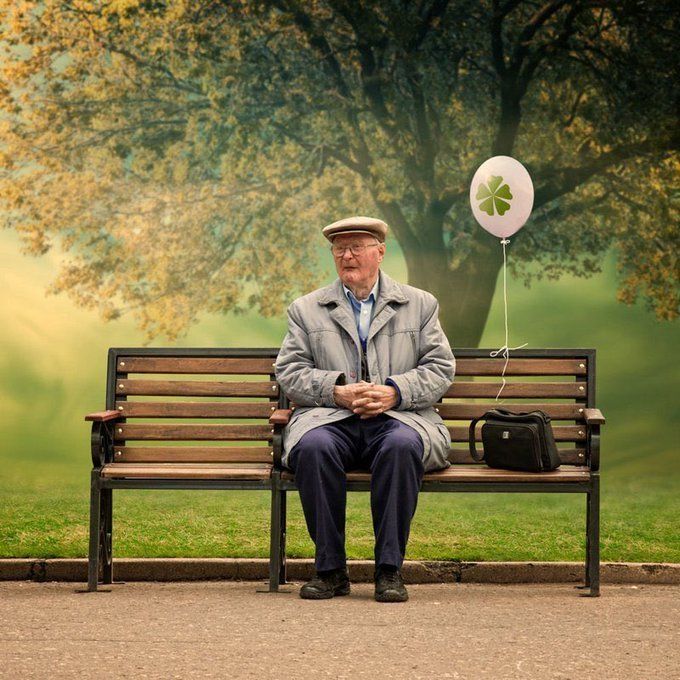 an old man sitting on a bench with a balloon attached to his hat and jacket