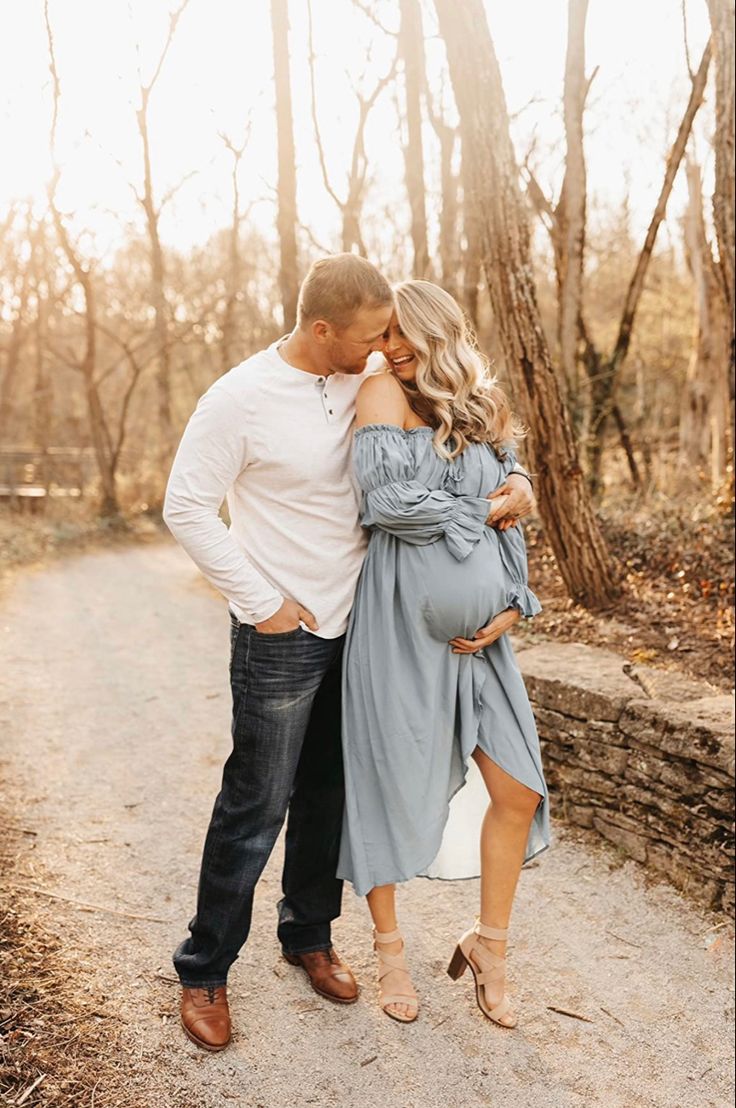 a pregnant couple kissing while standing on a dirt road in the woods with trees behind them