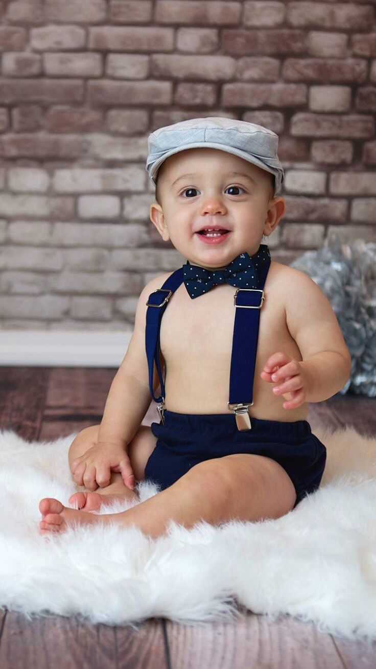 a baby wearing a hat and suspenders is sitting on a white fur rug in front of a brick wall