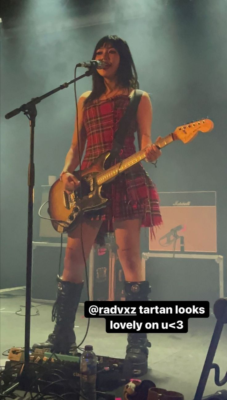 a woman in plaid dress holding a guitar on stage with microphone and keyboard behind her