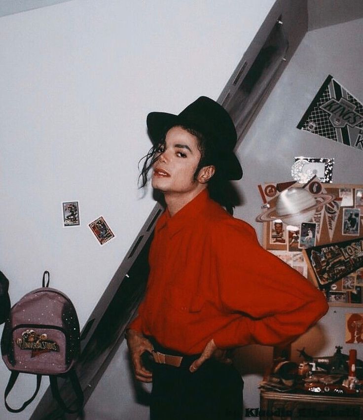 a man in a red shirt and black hat standing next to a wall with pictures on it