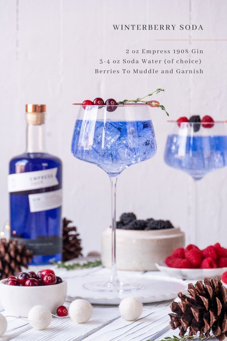 two glasses filled with blue liquid sitting on top of a table next to pine cones and berries