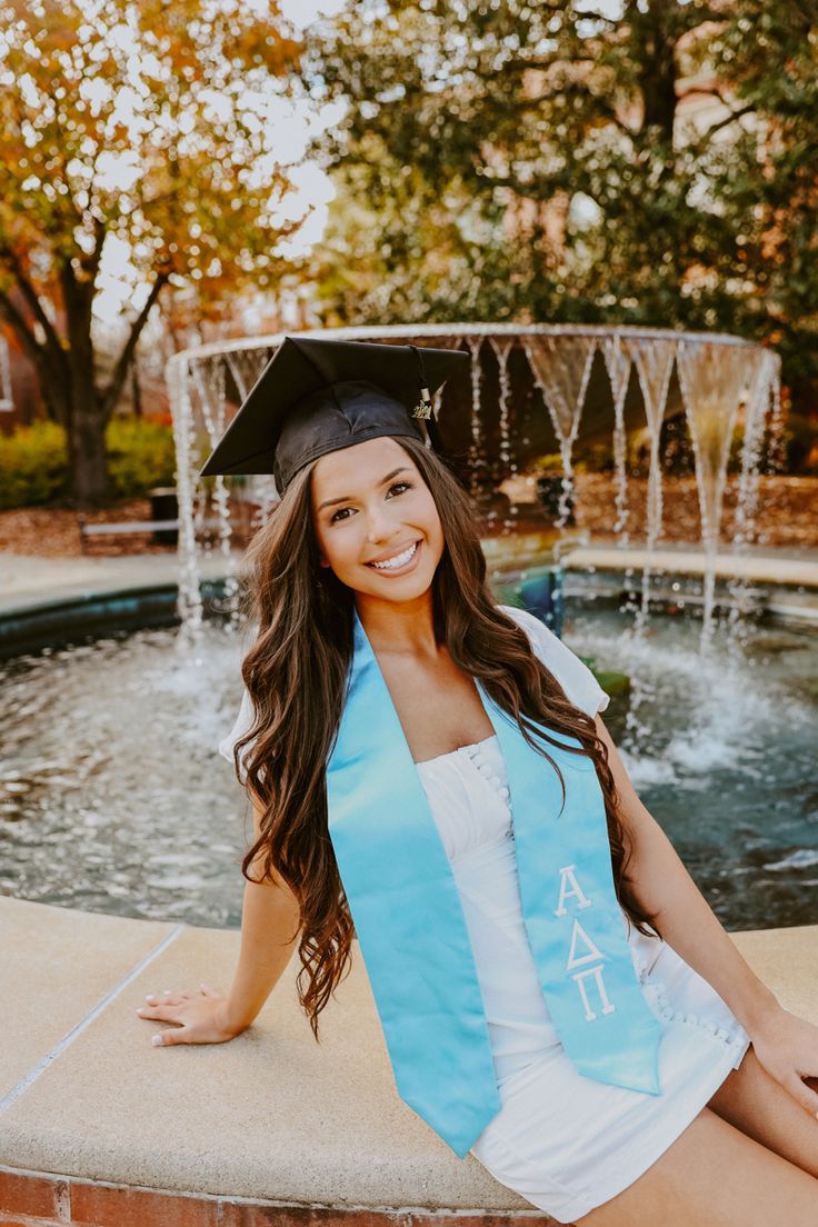 a woman sitting on the edge of a fountain wearing a graduation cap and blue sash