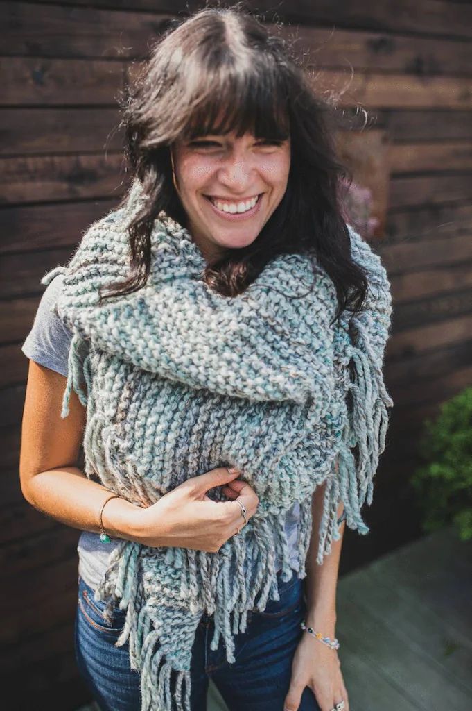 a smiling woman wearing a knitted shawl with fringes on the bottom and sides