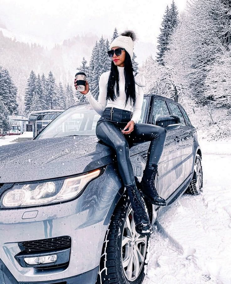 a woman sitting on top of a car in the snow holding a cup of coffee