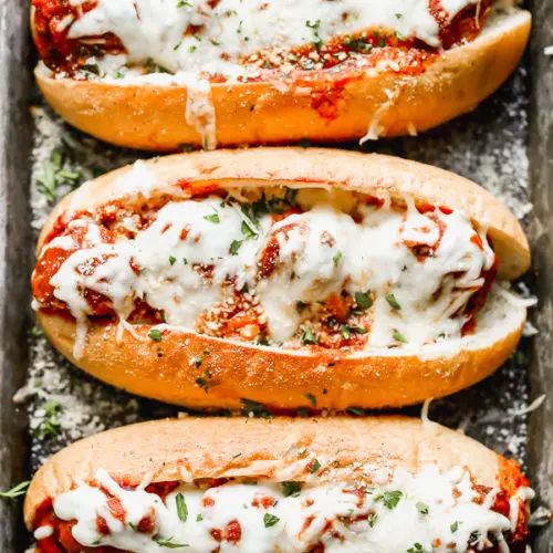 three hot dogs in buns covered with cheese and sauce on a baking sheet, ready to be eaten