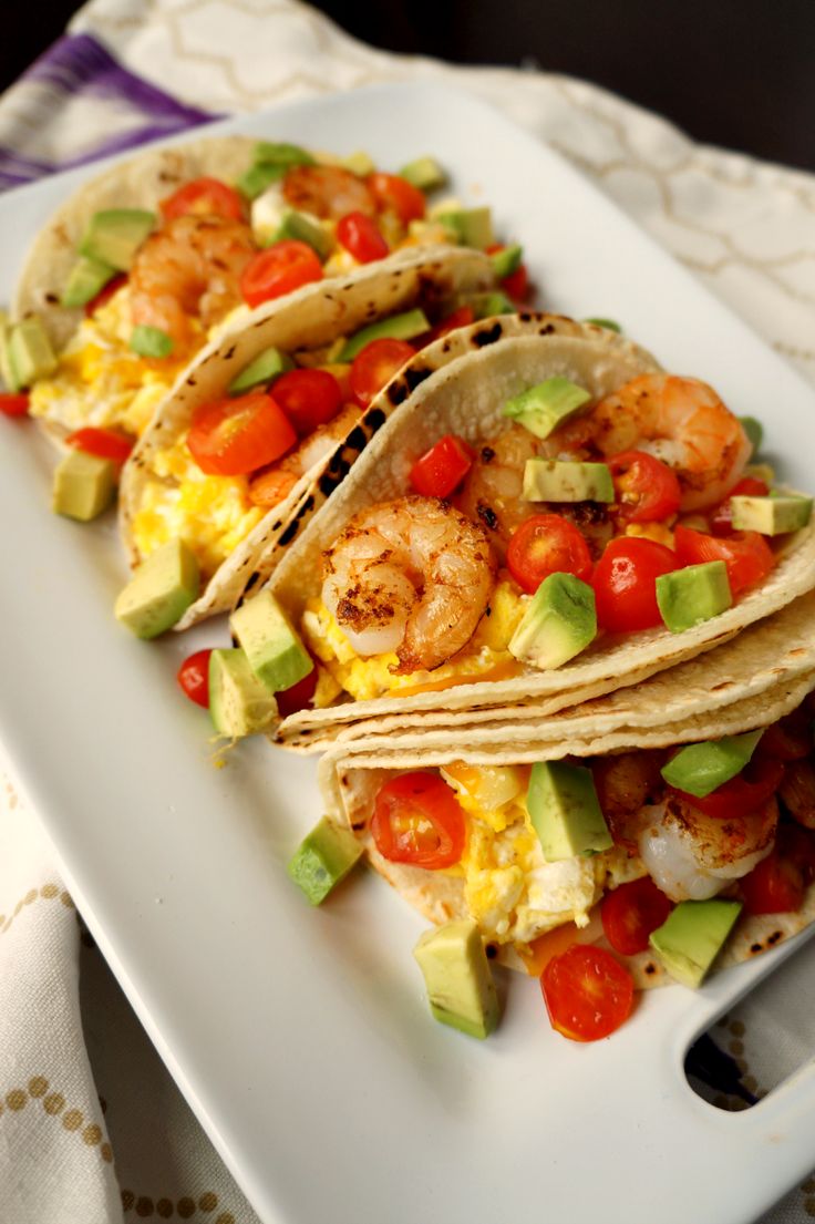 three tacos with shrimp, avocado and tomatoes on a white platter