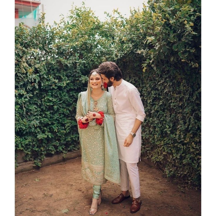 a man and woman standing next to each other in front of some bushes with flowers