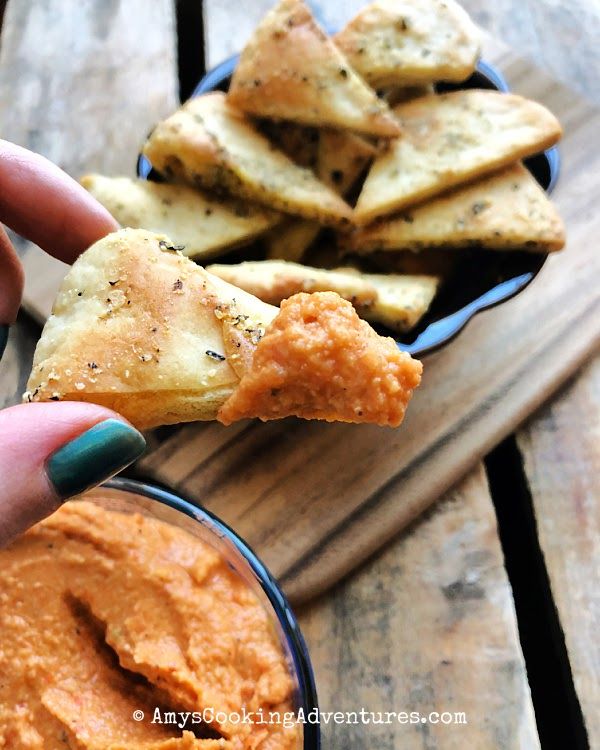 a hand holding up a piece of bread with dipping sauce in front of some pita chips