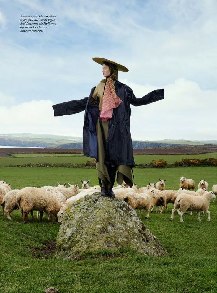 a woman standing on top of a rock in front of a herd of sheep and wearing a hat