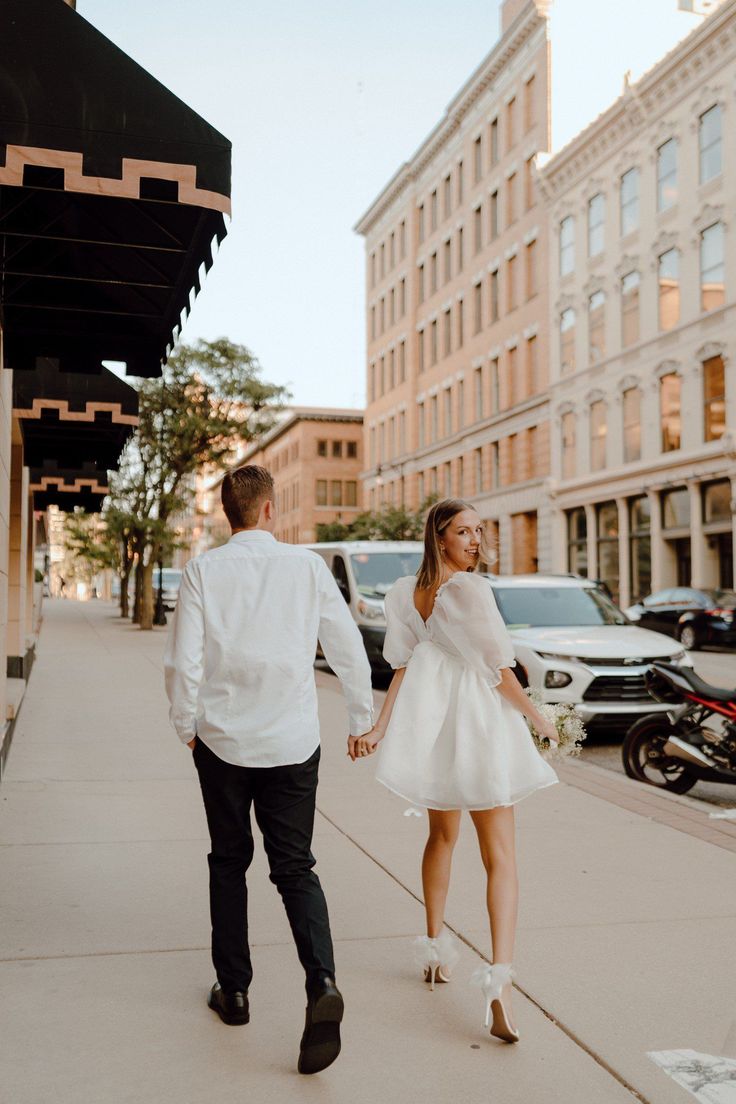 a man and woman are walking down the sidewalk in front of tall buildings, holding hands