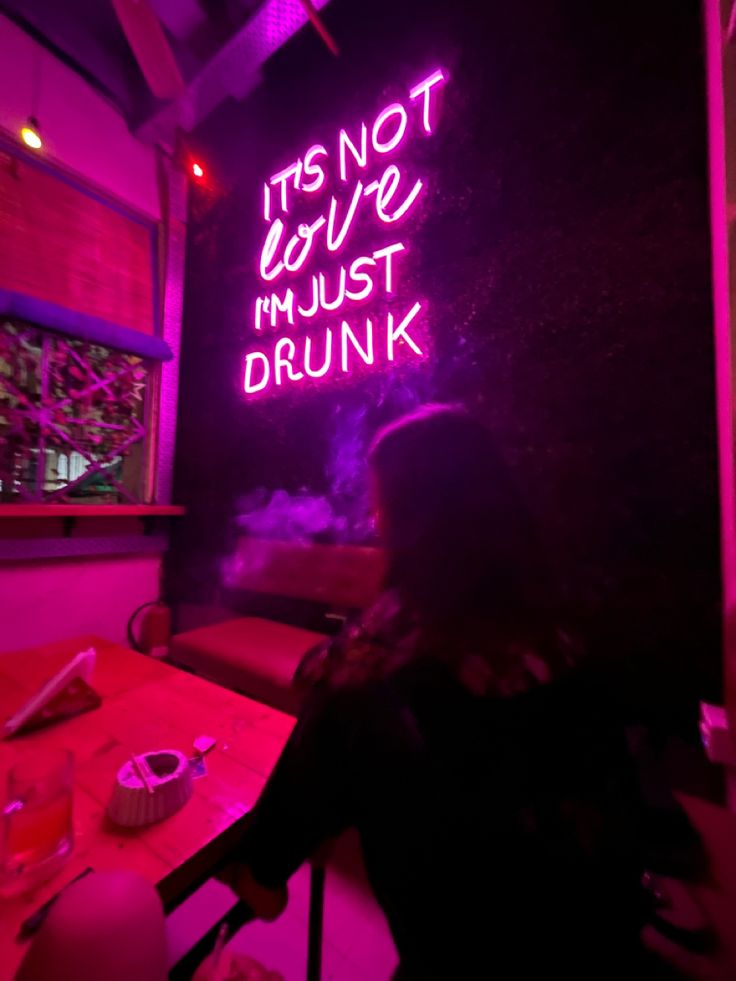 a woman sitting at a table in front of a neon sign that says it's not love must drunk