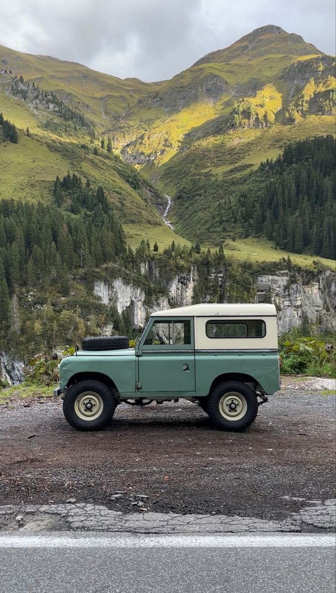 an old green truck parked on the side of a road in front of some mountains