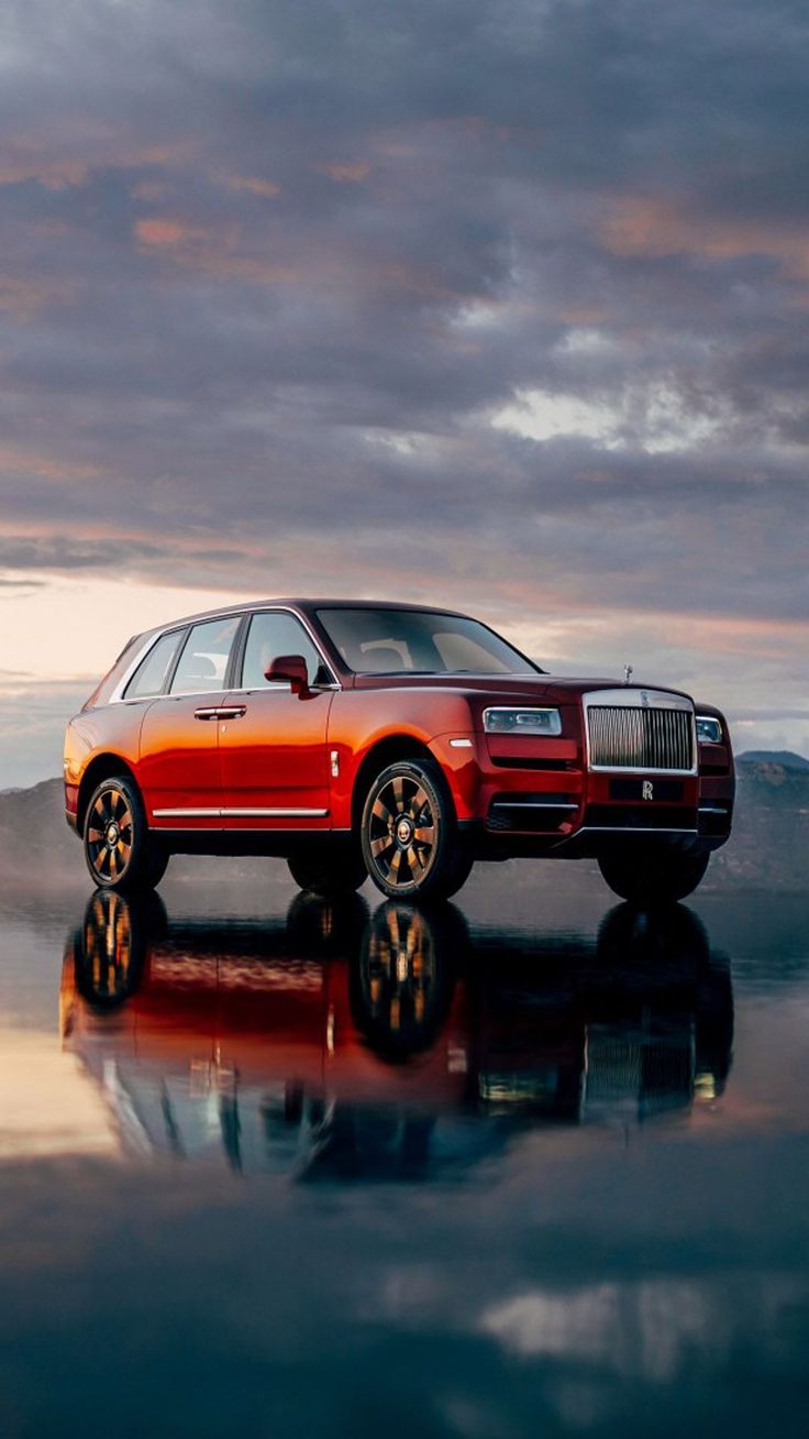 a red car parked in front of a body of water with clouds above it and reflecting on the ground