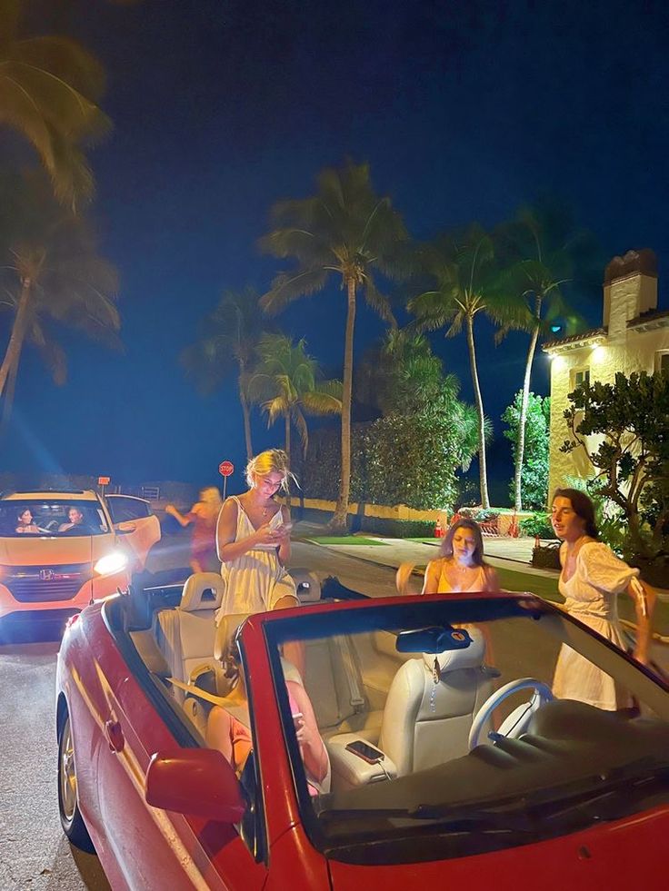 two women sitting in the back of a red convertible car at night with palm trees behind them