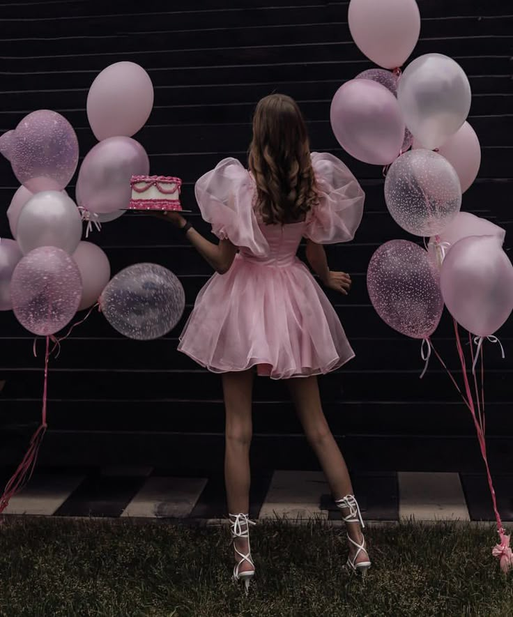 a girl in a pink dress holding a piece of cake with balloons around her and the back of her head