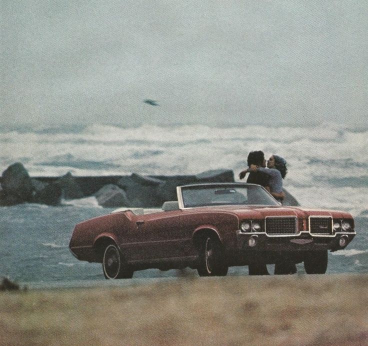 two people are sitting on the roof of a convertible car near the ocean and rocks