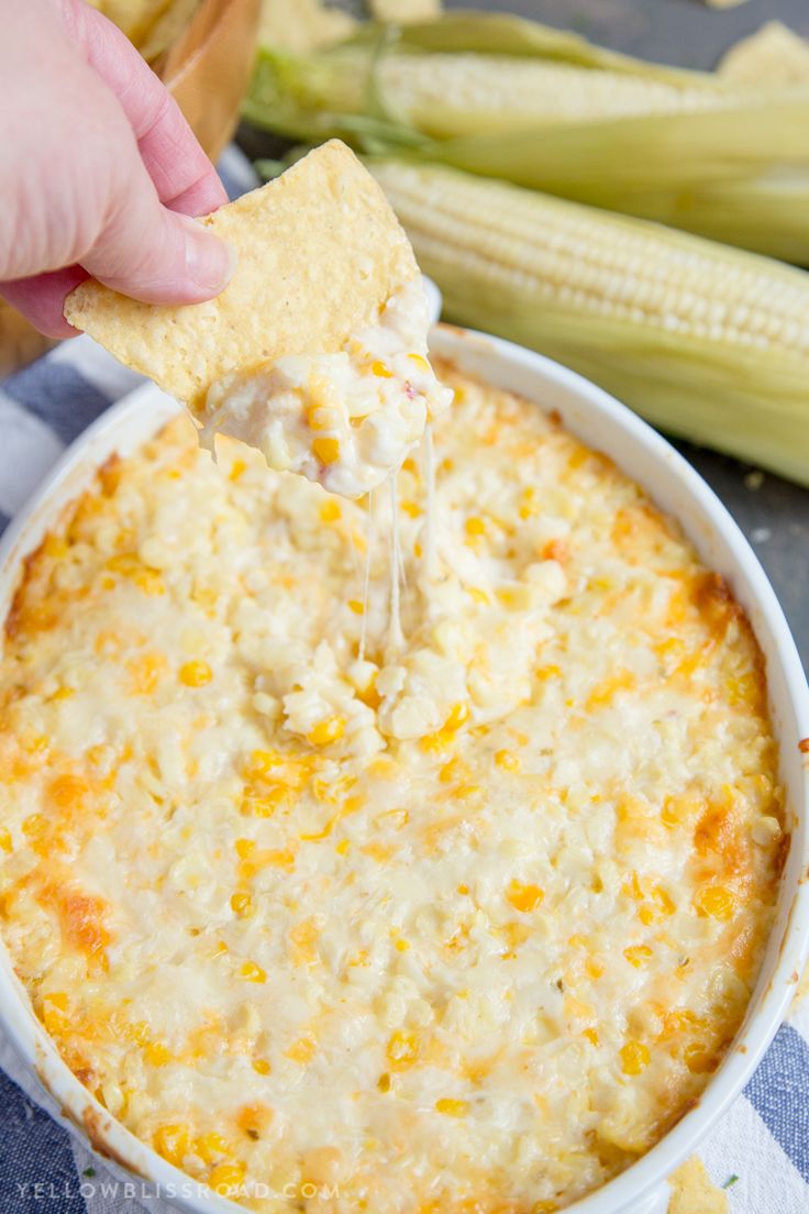 a hand holding a tortilla chip over a bowl of cheesy corn dip