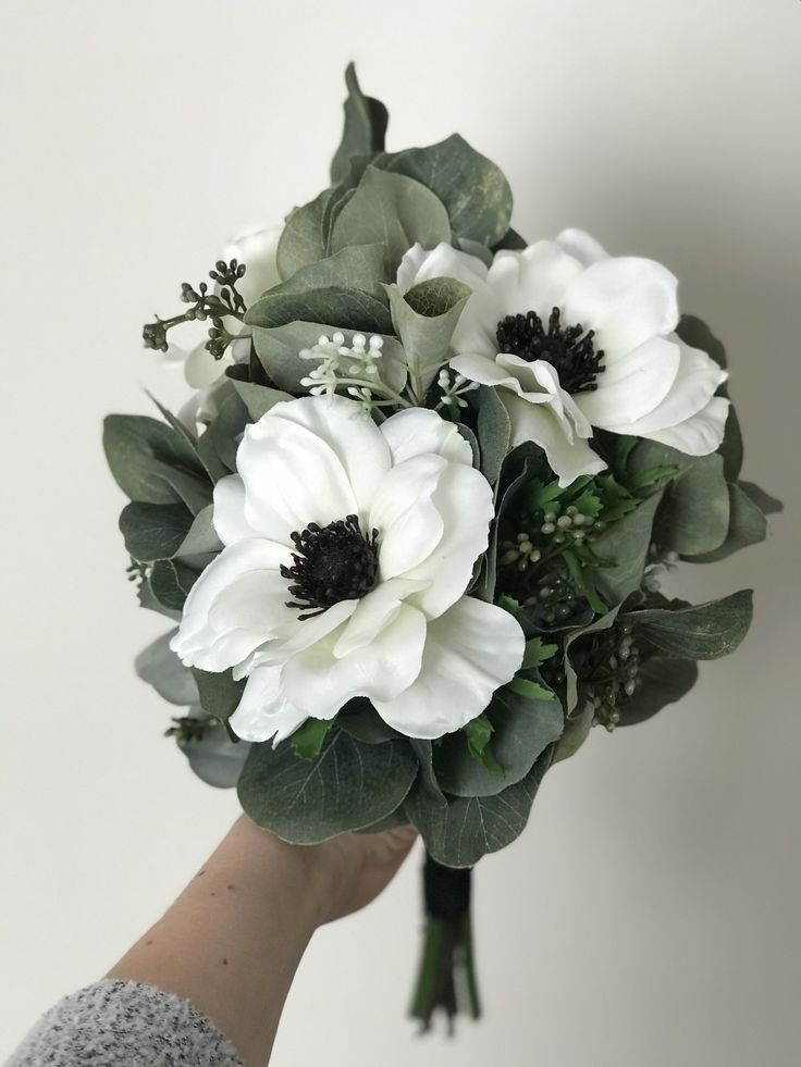 a person holding a bouquet of white flowers in their hand with greenery on it