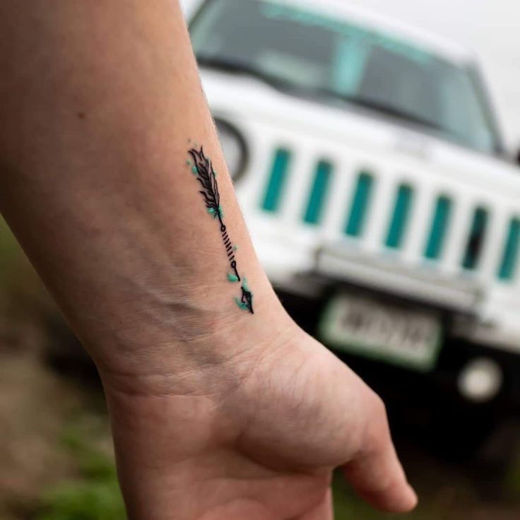 a small wrist tattoo with an arrow on the left side of the arm and a jeep in the background