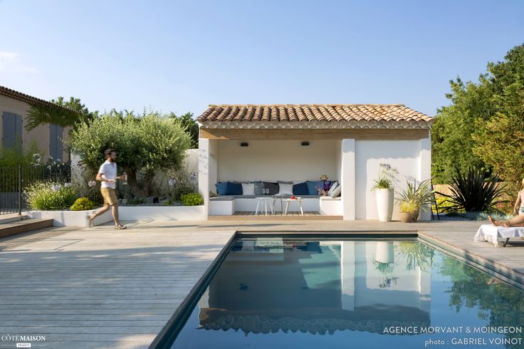 two people are sitting by the pool in front of a house with an outdoor dining area