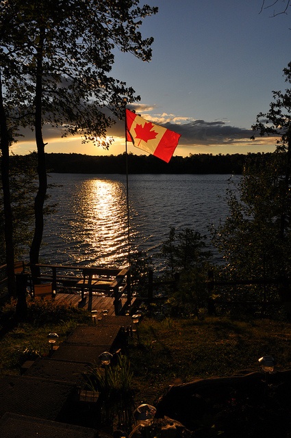 the canadian flag is flying high over the water at sunset or dawn, with trees lining the shore