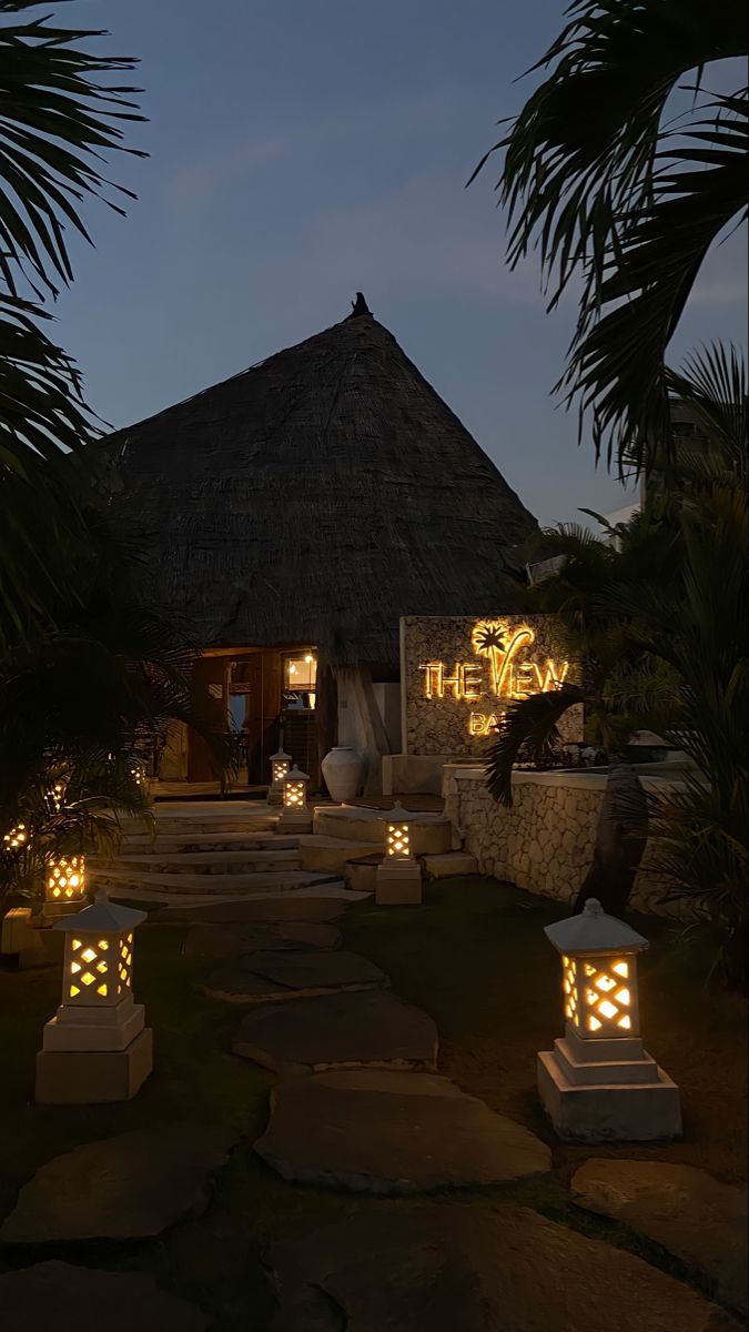 some lights are lit up in front of a hut at night with palm trees and steps leading to it