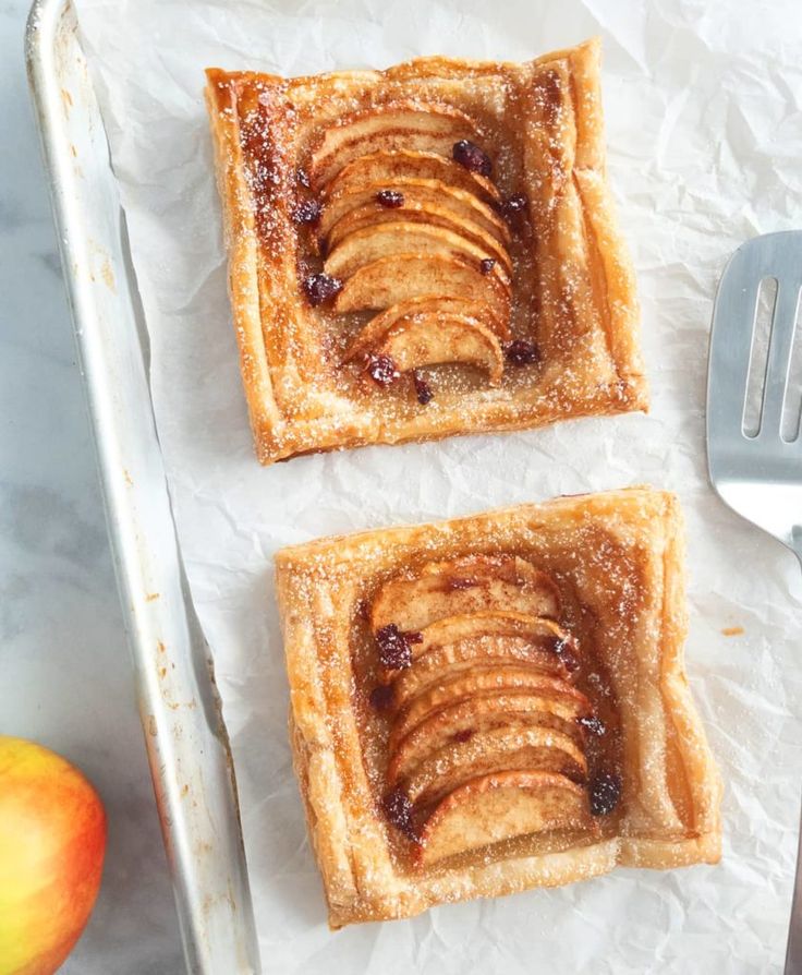 two slices of apple pie on top of parchment paper next to an apple and fork