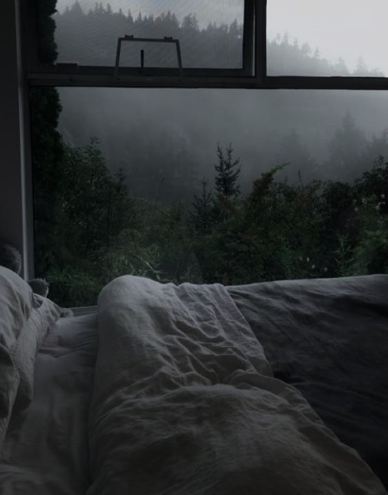 an unmade bed in front of a window with foggy trees outside it and a chair on the other side