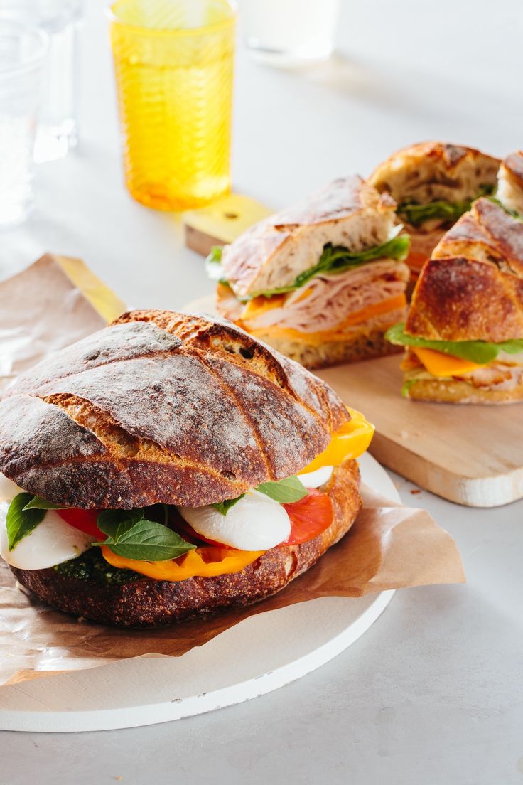 two sandwiches with meat, cheese and veggies are on wooden cutting boards next to each other