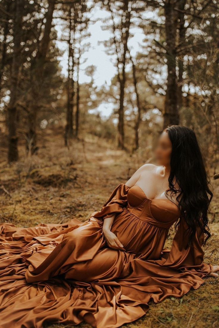 a woman in a brown dress sitting on the ground next to some trees and grass