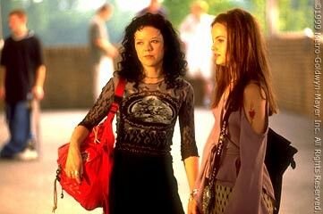 two women standing next to each other on a sidewalk with people in the background and one woman carrying a red purse