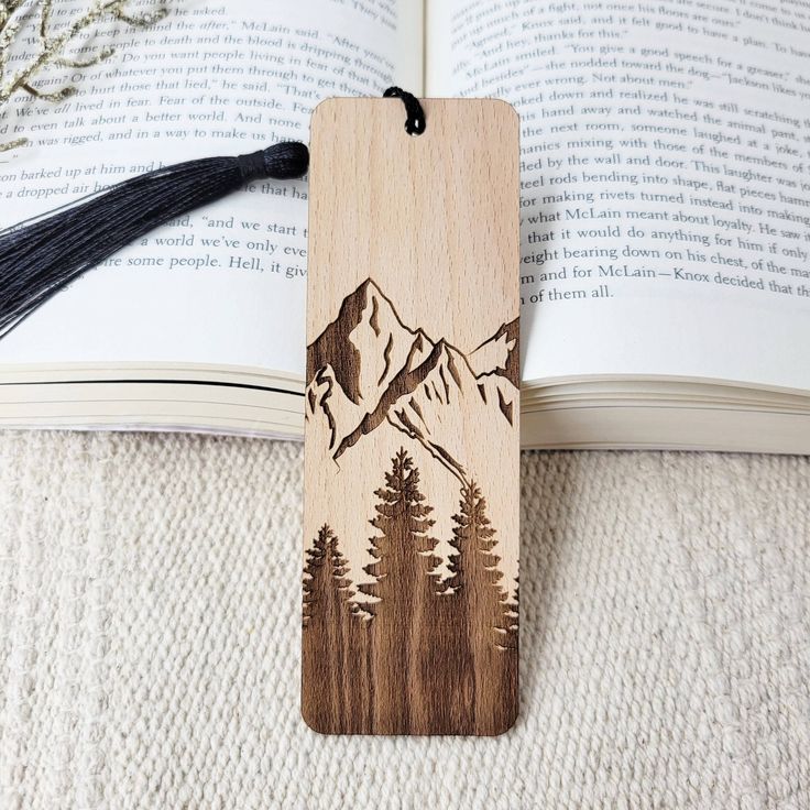 a wooden bookmark with mountains and trees on it sitting next to an open book