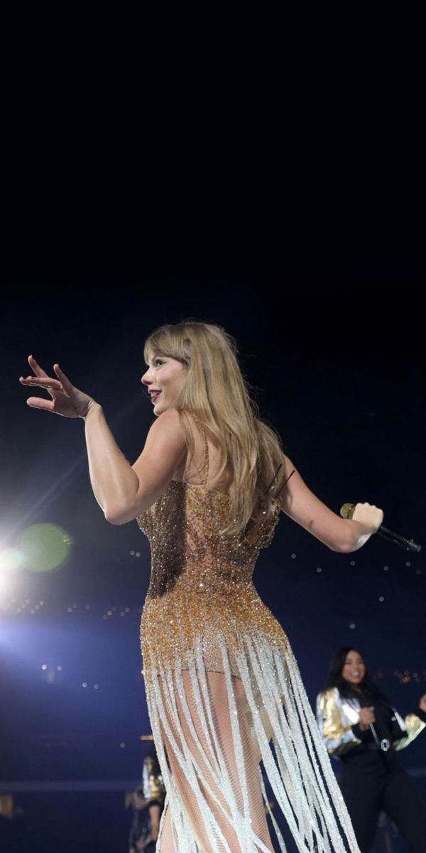 a woman in a gold and white dress on stage with her arms out to the side