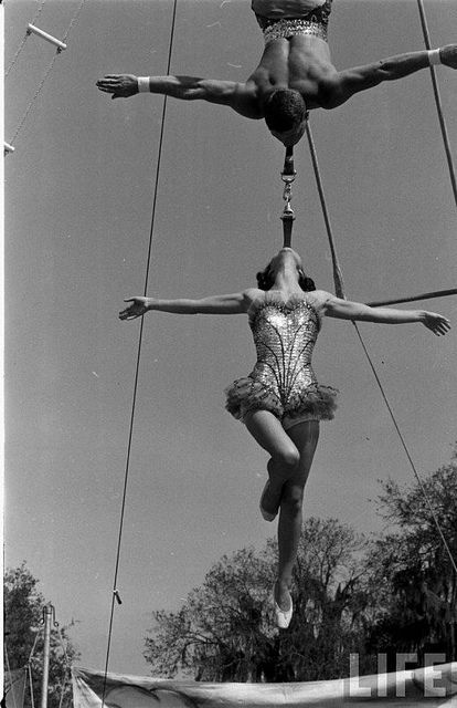 two acrobatic performers performing aerial tricks in an outdoor circus ring, mid - 1960s