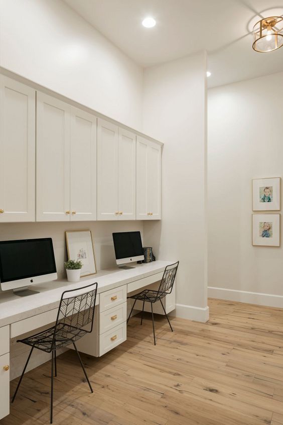 two computer desks sitting on top of a wooden floor next to white cupboards