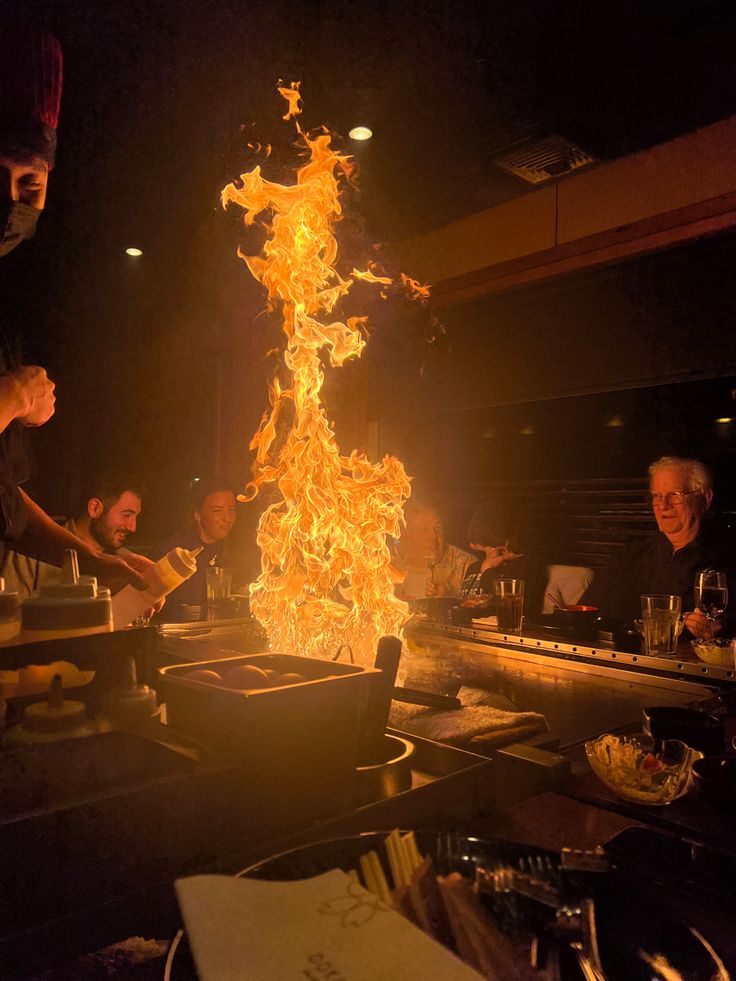 a group of people sitting around a table with food on it and flames in the air