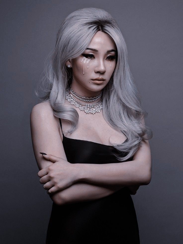 a woman with long blonde hair wearing a black dress and diamond choker, standing in front of a gray background