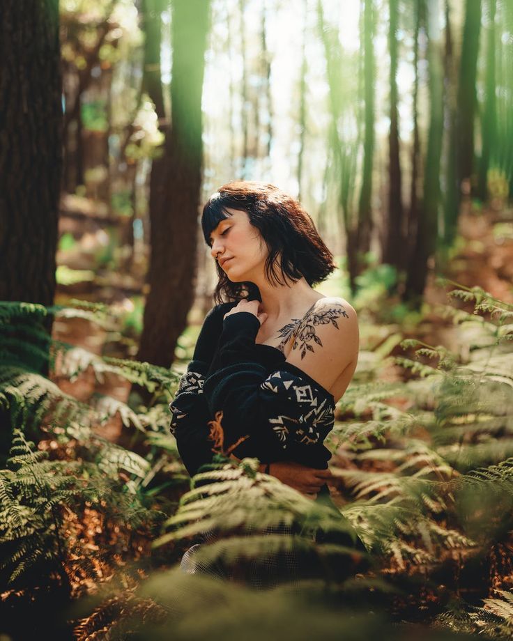 a woman standing in the middle of a forest surrounded by tall trees and ferns, looking off into the distance