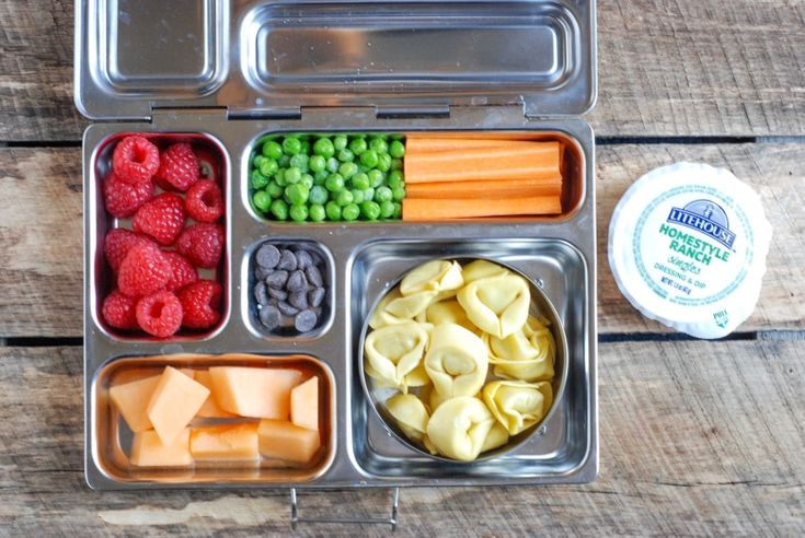 an open lunch box with fruits, vegetables and pasta in it on a wooden table