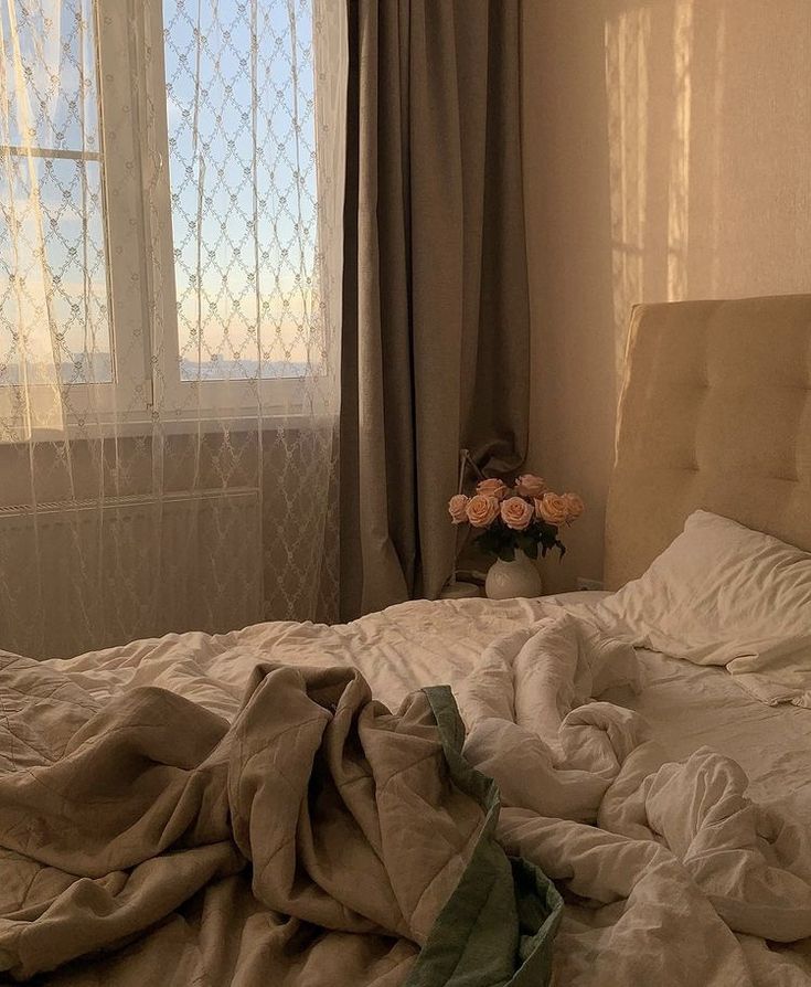 an unmade bed in front of a window with curtains and flowers on the side