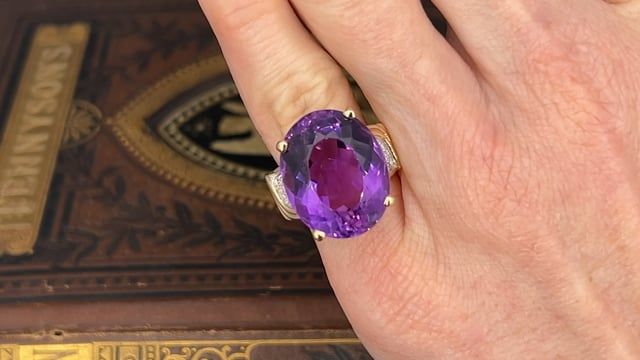 This amethyst cocktail ring is centered with one (1) oval mixed cut natural amethyst set into a four-prong setting. The shoulders of the ring are each accented with five (5), bead set, round brilliant cut diamonds. The ring measures 20.0mm at the top, rises 12.1mm above the finger, tapering to 3.3mm wide and 0.9mm thick at the base of the shank. The ring is currently a size 5. Luxury Purple Oval Gemstones, Oval Amethyst Ring With Gemstone Accents For Formal Occasions, Oval Amethyst Gemstones For Formal Occasions, Formal Oval Amethyst Gemstones, Heirloom Style Purple Oval Amethyst Ring, Oval Amethyst Ring For Formal Occasions, Formal Oval Amethyst Ring In Fine Jewelry Style, Oval Purple Amethyst Ring With Gemstone Accents, Oval Amethyst Ring With Center Stone For Formal Occasions