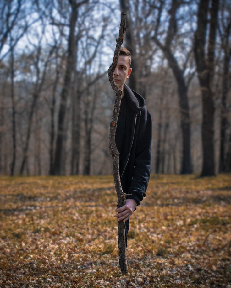 a young man holding onto a tree branch in the middle of a forest with leaves on the ground