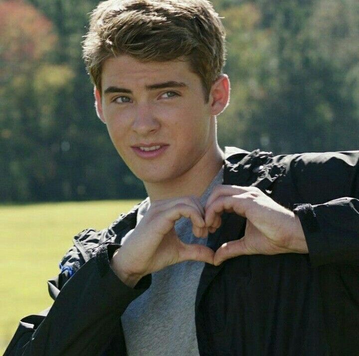 a young man making a heart shape with his hands while standing in front of a field