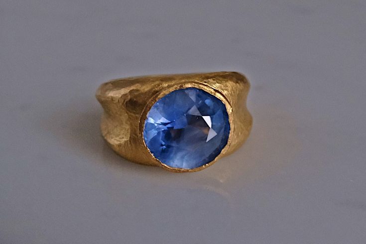 Fine 24k gold ring set with an unheated Sri Lankan sapphire weighing 7.5ct. The round blue sapphire is light cornflower blue. The stone has medium saturation, but its light cornflower hue is unique. This unique blue wrapped by the purity of gold sets up a ring with opposing lights. The stone is cool and the gold is warm - the ring carries a light stone, as well as a light-infused stone. In any hue of blue, a round sapphire is harder to find than one can imagine. The band is reminiscent of Roman rings, except an actual Roman ring will not be as hefty. The ring now weighs 21.69 grams, a size 7. This design is built on ideas of continuity, the round stone flowing to its sculpted gold work. At this stone's size, its design will take any finger size, it is quintessentially Zen. The stone measur Yellow Gold Sapphire Ring With Faceted Detail, Blue Faceted Sapphire Ring Fine Jewelry, Blue Faceted Sapphire Ring In Fine Jewelry Style, Blue Faceted Sapphire Ring For Wedding, Formal Faceted Blue Sapphire Ring, Formal Blue Faceted Sapphire Ring, Blue Faceted Topaz Ring For Anniversary, Anniversary Blue Faceted Topaz Ring, Faceted Yellow Gold Sapphire Ring