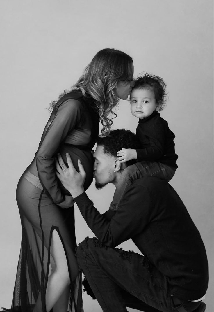 a man kneeling down next to a woman holding a baby and kissing her face on the cheek