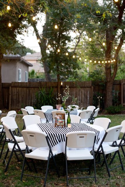 an outdoor party with white chairs and black and white table cloths, lights strung from the trees